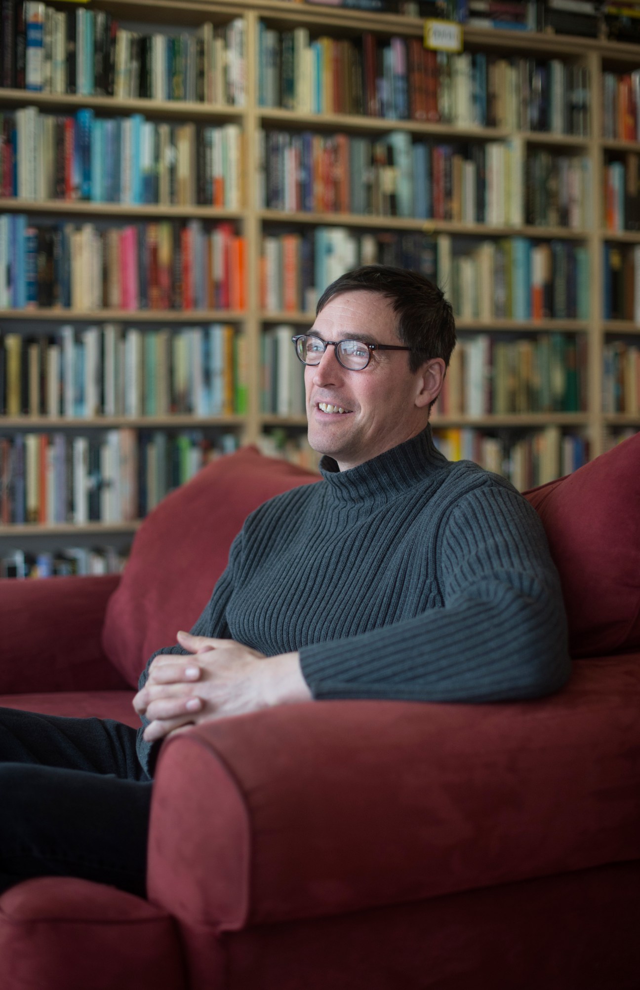 Man wearing glasses and dark sweater sits on burgundy armchair in front of bookshelves.