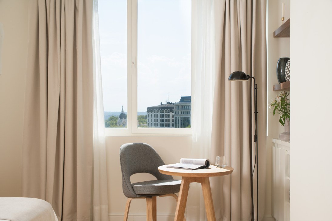 Hotel room with gray chair at wooden desk by window overlooking Vienna cityscape.
