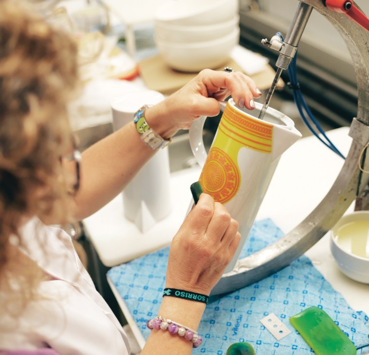 A craftsperson applies colorful decals to white porcelain using a specialized tool during the decorating process.