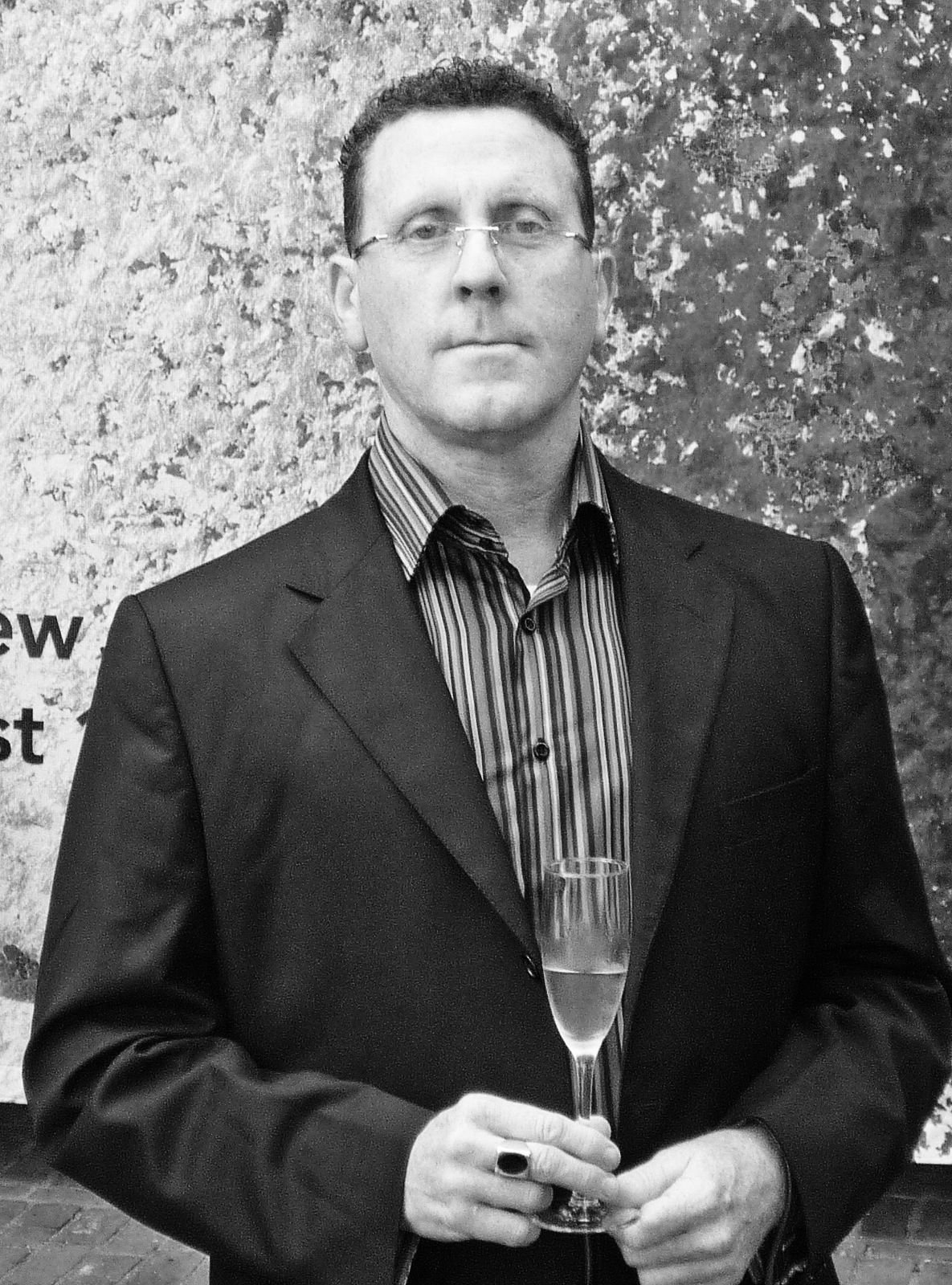 Black and white portrait of David Anfam, commissioning editor for fine art at Phaidon Press, holding a glass against a textured wall.