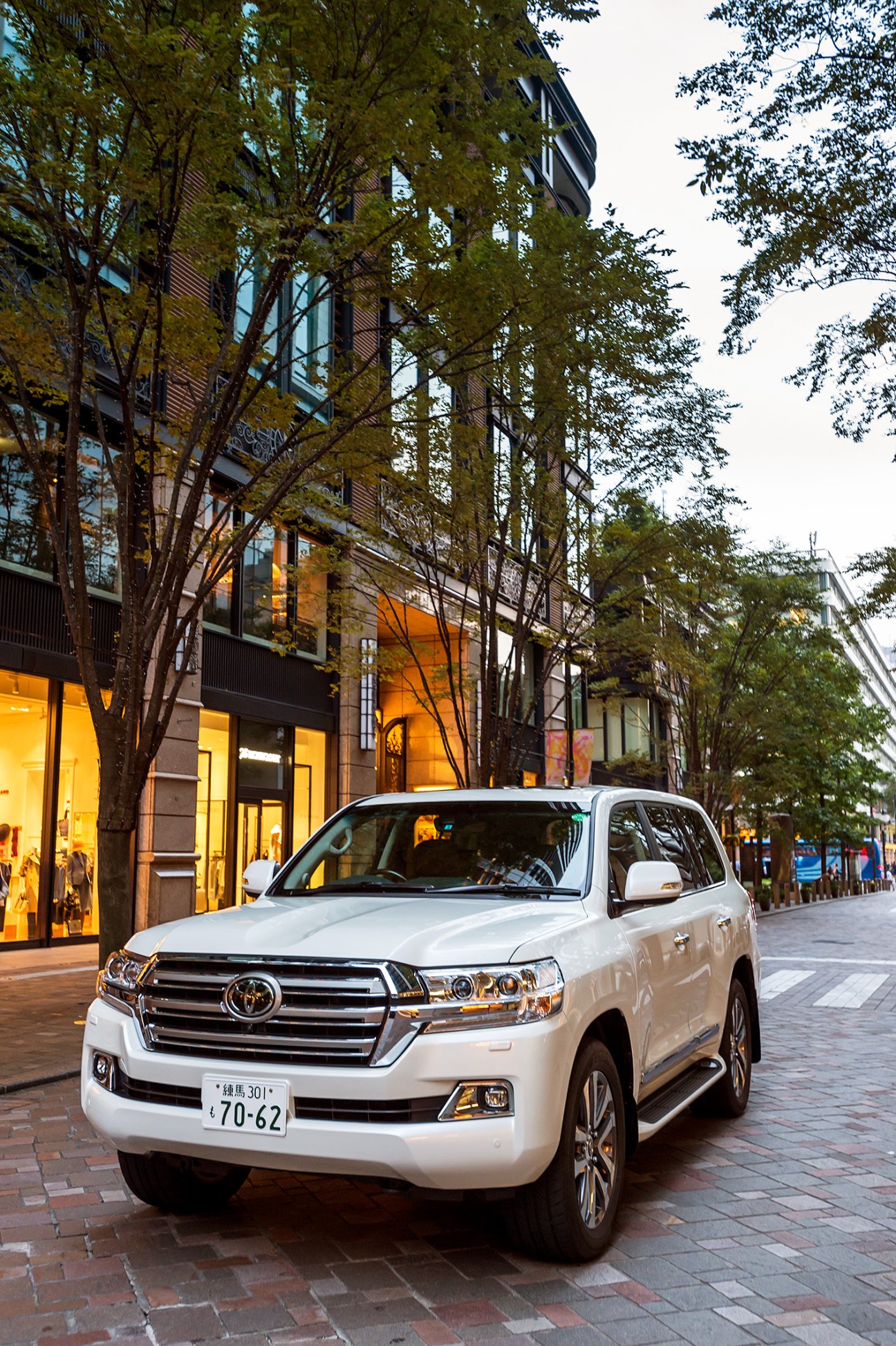White Toyota Land Cruiser SUV parked on a tree-lined urban street with illuminated storefronts.