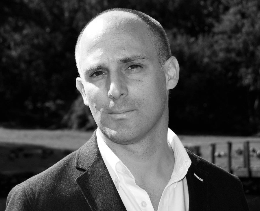 Black and white portrait photograph of a man in a dark jacket and white shirt against a blurred outdoor background.