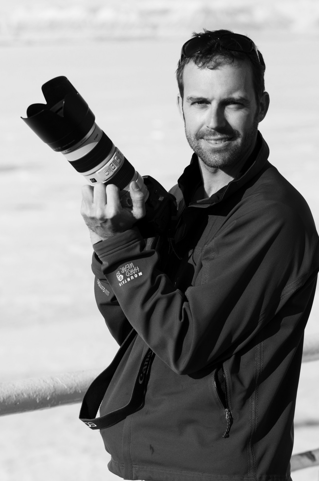 Filmmaker Ian Kellett holds a professional camera with telephoto lens while smiling at the camera in black and white.