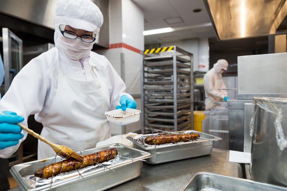 Chef in white uniform and hairnet preparing grilled skewers in an aircraft catering kitchen.