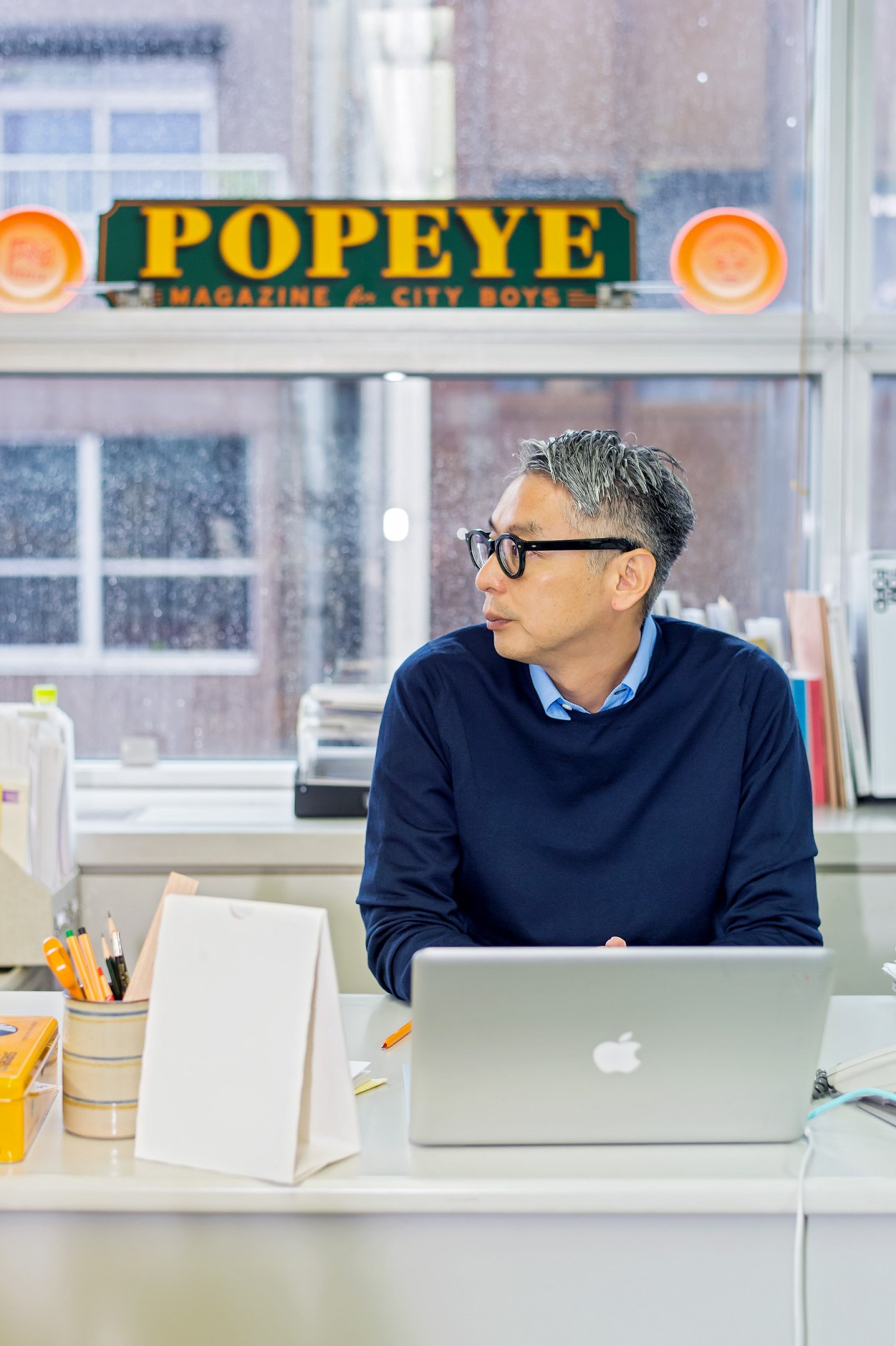 Man wearing glasses and navy sweater working at laptop in office with Popeye magazine sign visible through window.
