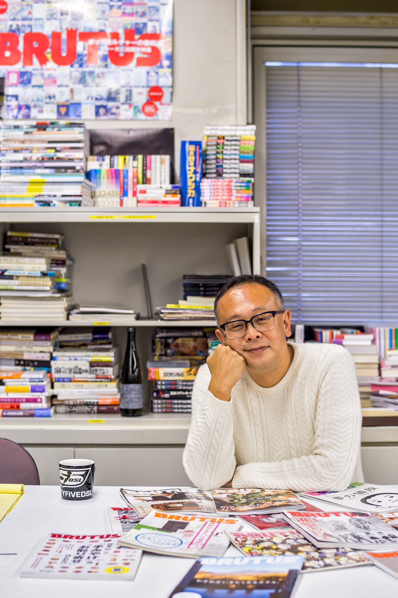 Tsutomu Ishizaki, president of Magazine House, sits at his desk surrounded by magazines and books with a Brutus poster visible behind him.