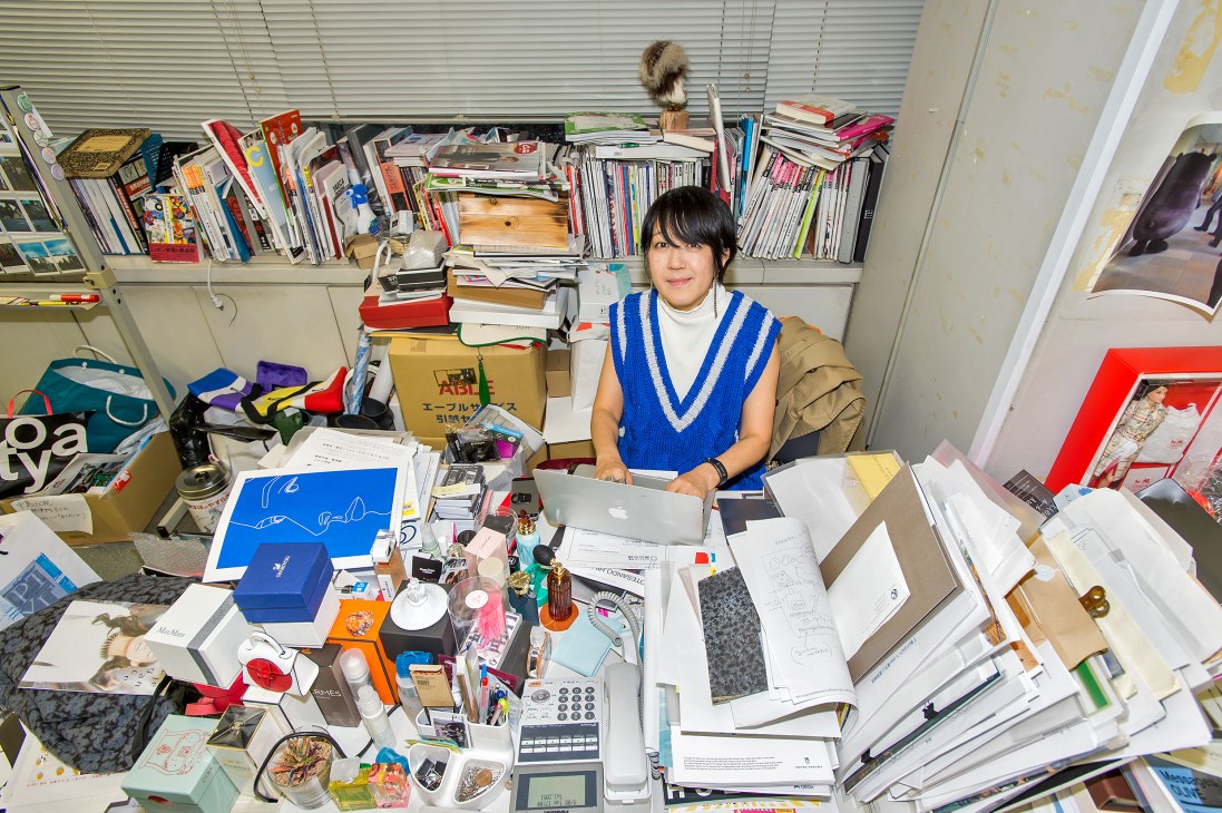 Toshiko Nakashima, editor in chief of Ginza magazine, sits at her cluttered desk surrounded by magazines, design materials, and office supplies.