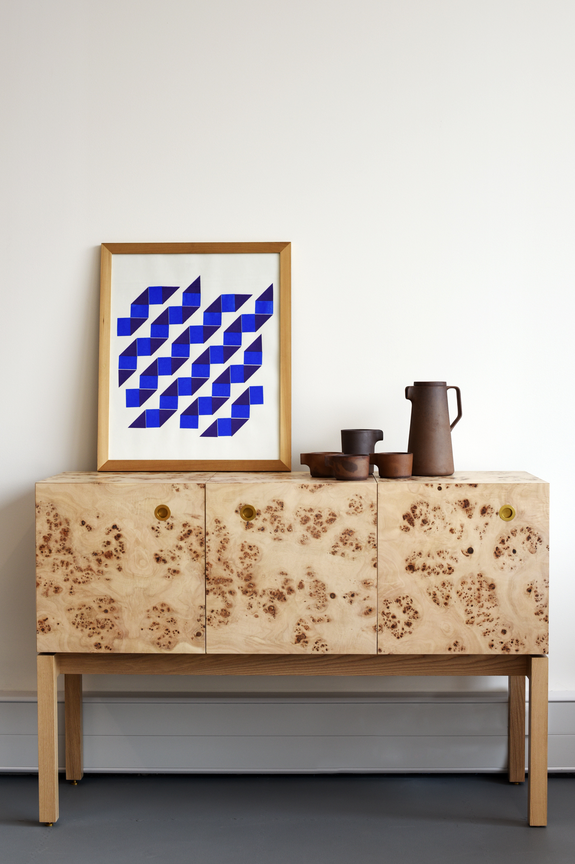 Wooden sideboard with burled veneer doors and ceramic vessels displayed on top next to framed geometric artwork.