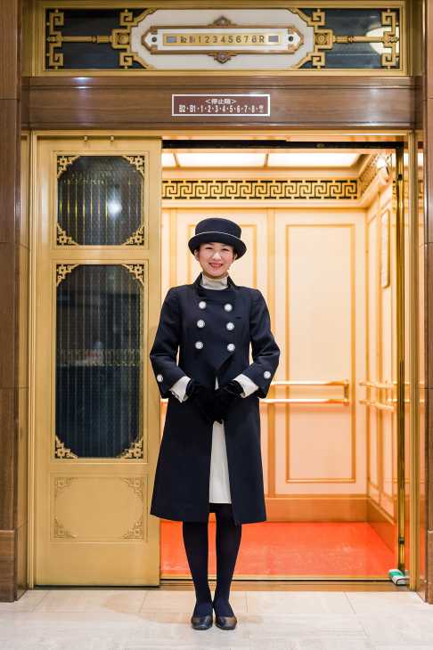 An elevator attendant in a black and cream uniform stands in a vintage gold-trimmed lift at Takashimaya, Tokyo.