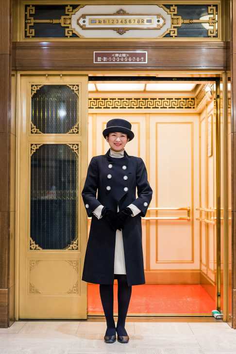 An elevator attendant in a black and cream uniform stands in a vintage gold-trimmed lift at Takashimaya, Tokyo.