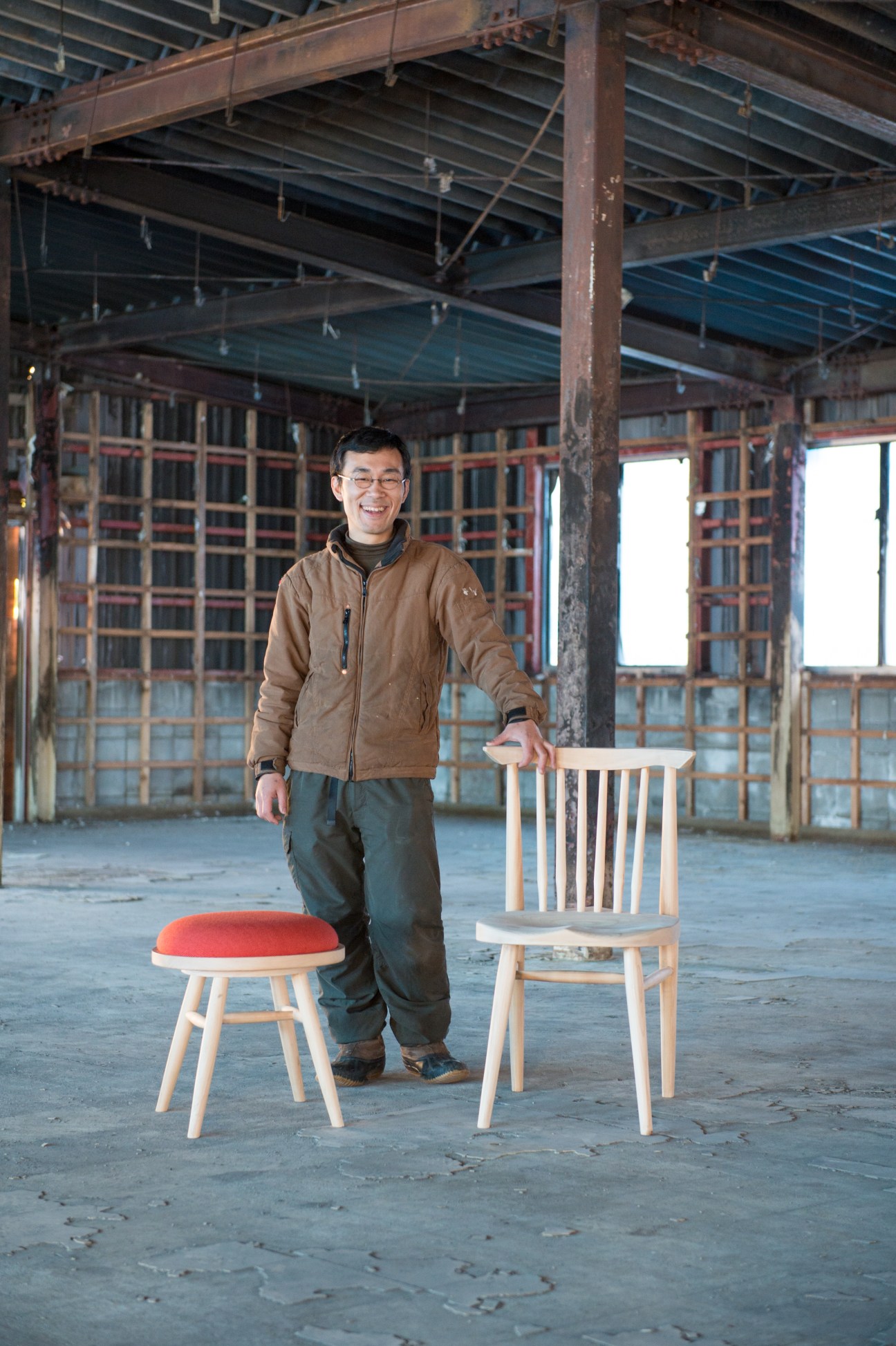 Man standing in an industrial space with wooden chairs, one with a red seat cushion.