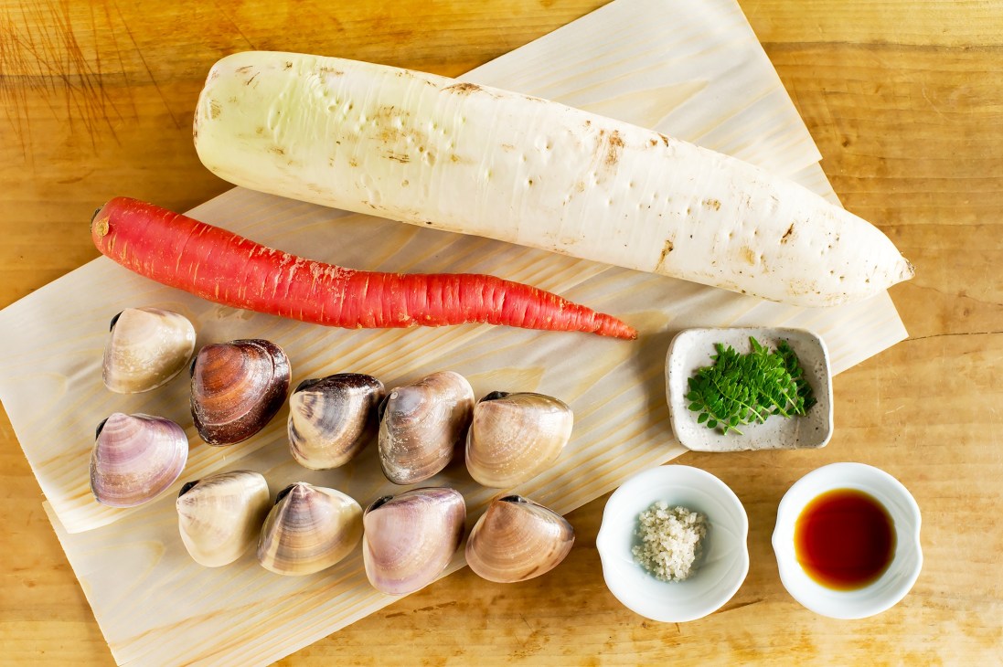 Ingredients for clam soup including clams, white radish, carrot, pepper leaves, and condiments arranged on wooden surface.