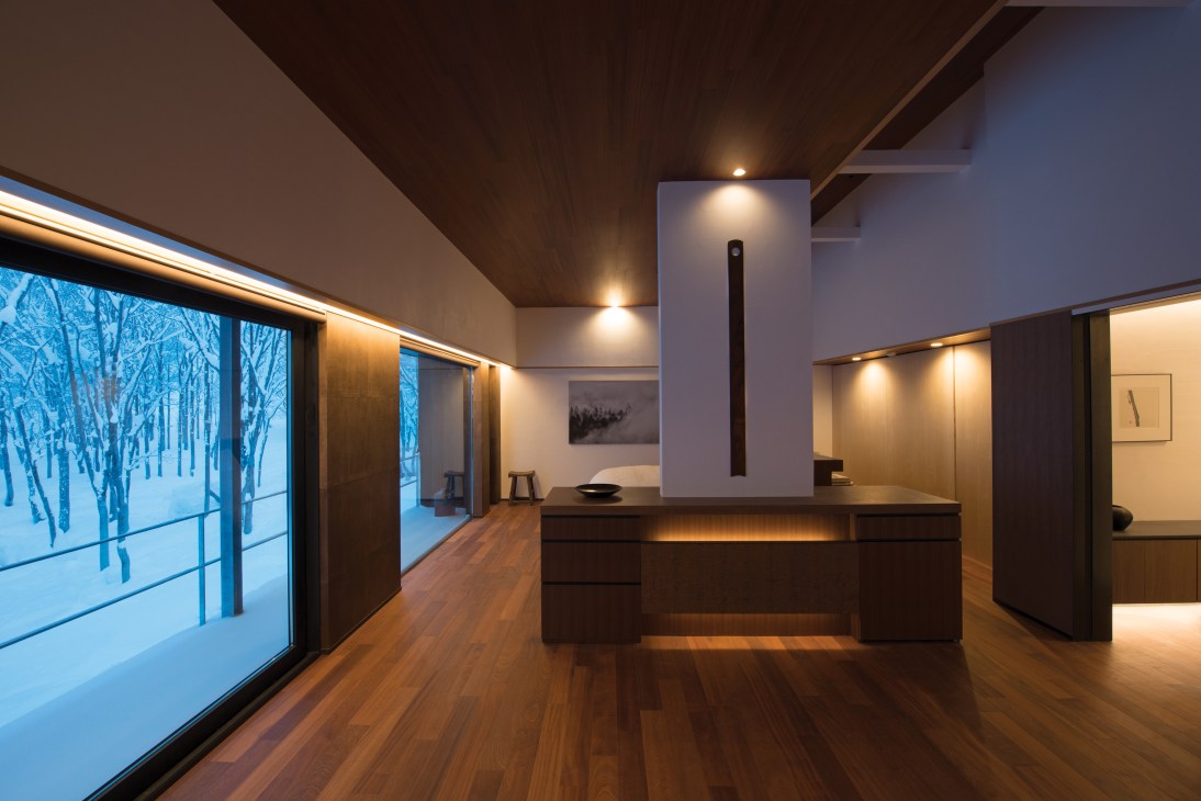 Modern interior of a luxury ryokan villa with wooden flooring, large windows overlooking snowy forest, and minimalist design with warm lighting.
