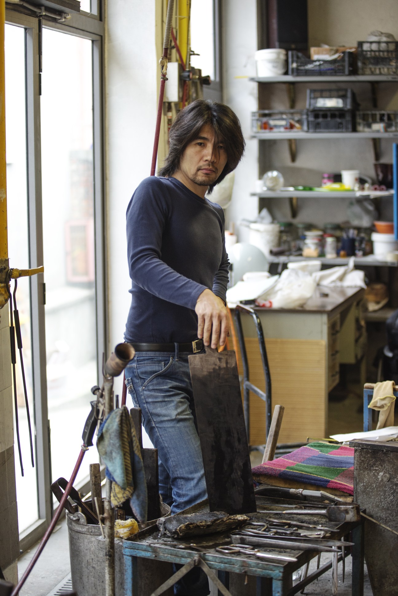 Yasuhiko Tsuchida, a Japanese glassmaker, stands in his Murano studio surrounded by glass-making tools and materials.