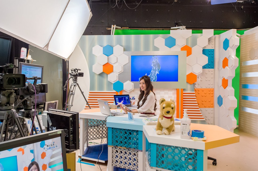 Weather forecast studio with colorful hexagonal wall panels, presenter at desk with laptops and screens displaying weather data.