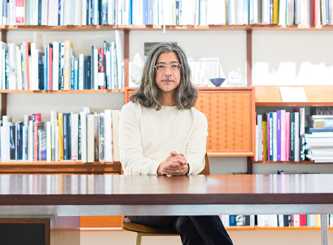 Woman with glasses and shoulder-length hair sits at wooden desk in front of floor-to-ceiling bookshelves