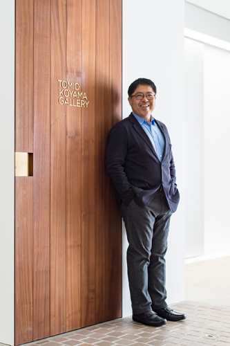 A smiling man in a dark suit stands next to wooden doors marked 'Tomio Koyama Gallery' in a modern white interior