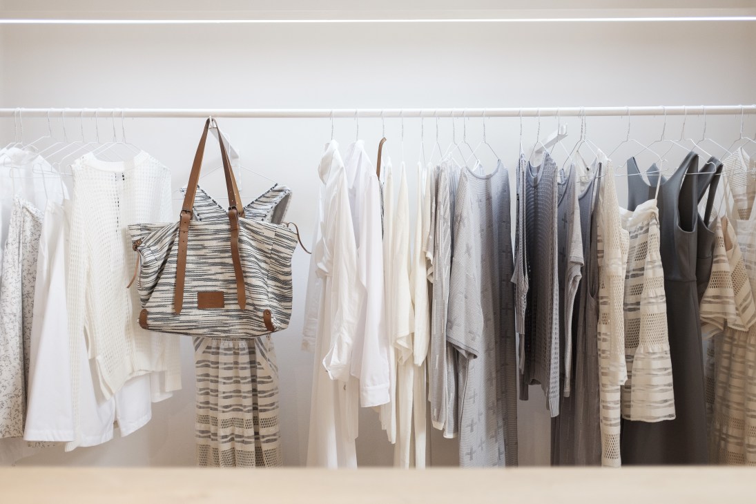 Clothing rack displaying white, cream, grey and black garments with striped tote bag hanging prominently.