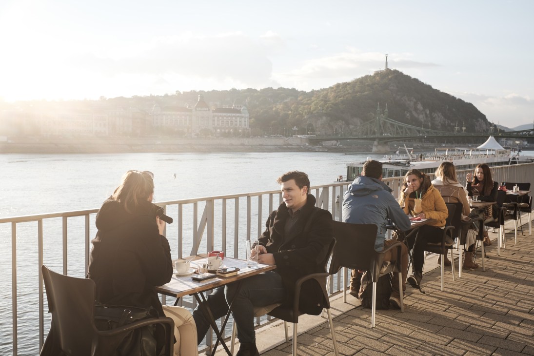 People dining on a riverside terrace overlooking the Danube with Budapest landmarks and a forested hill in the background.