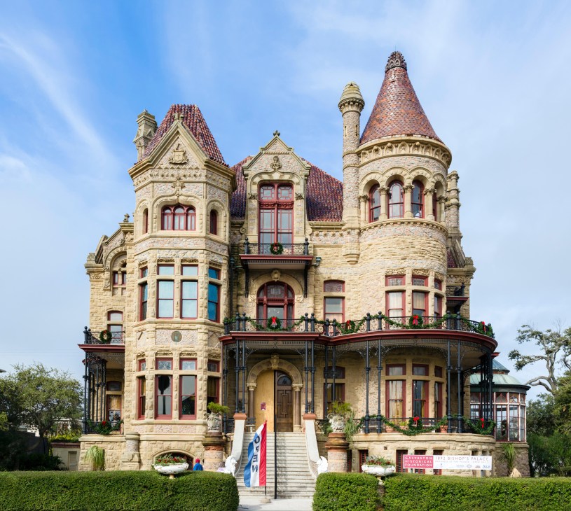 Victorian mansion with ornate stone facade, turrets and red-tiled roofs against blue sky.