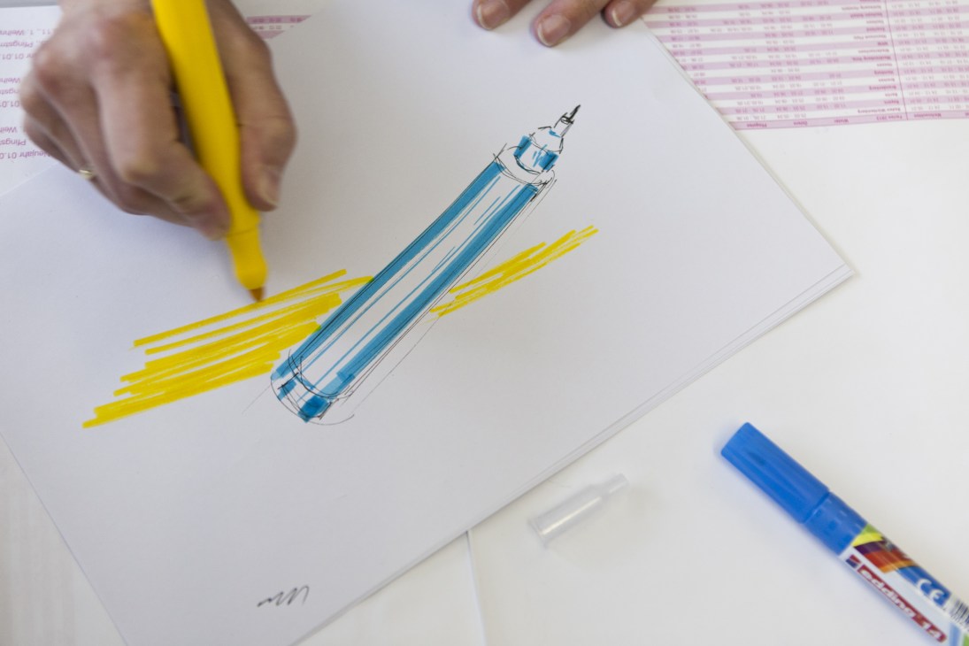 Hand holding a yellow Edding marker drawing lines next to a pen sketch on white paper.