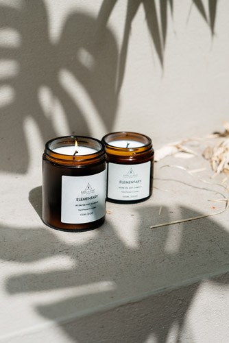 Two scented candles in amber glass jars with white labels on a white surface with plant shadows.