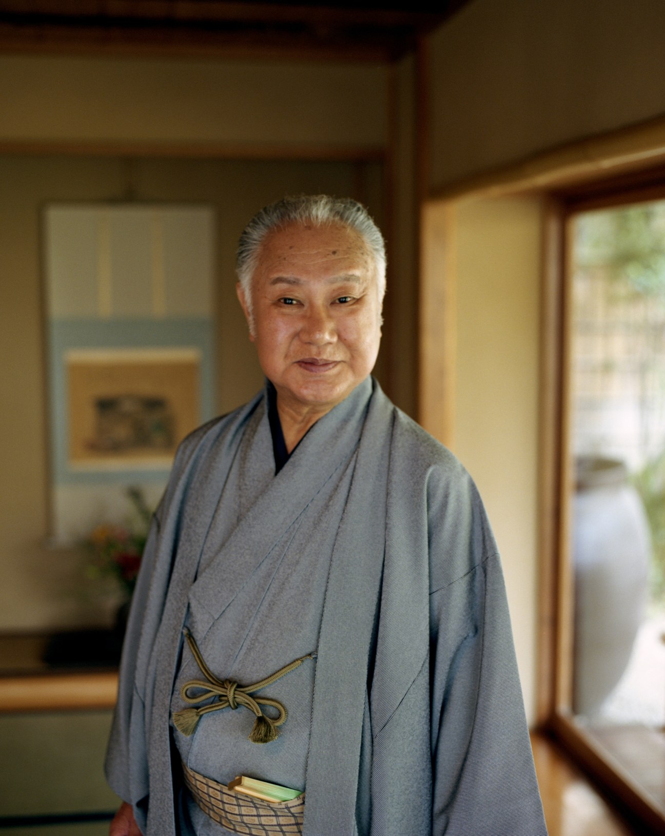 Tojuro Sakata, an elderly Japanese kabuki actor wearing a grey kimono with decorative gold obi, standing in a traditional Japanese interior.
