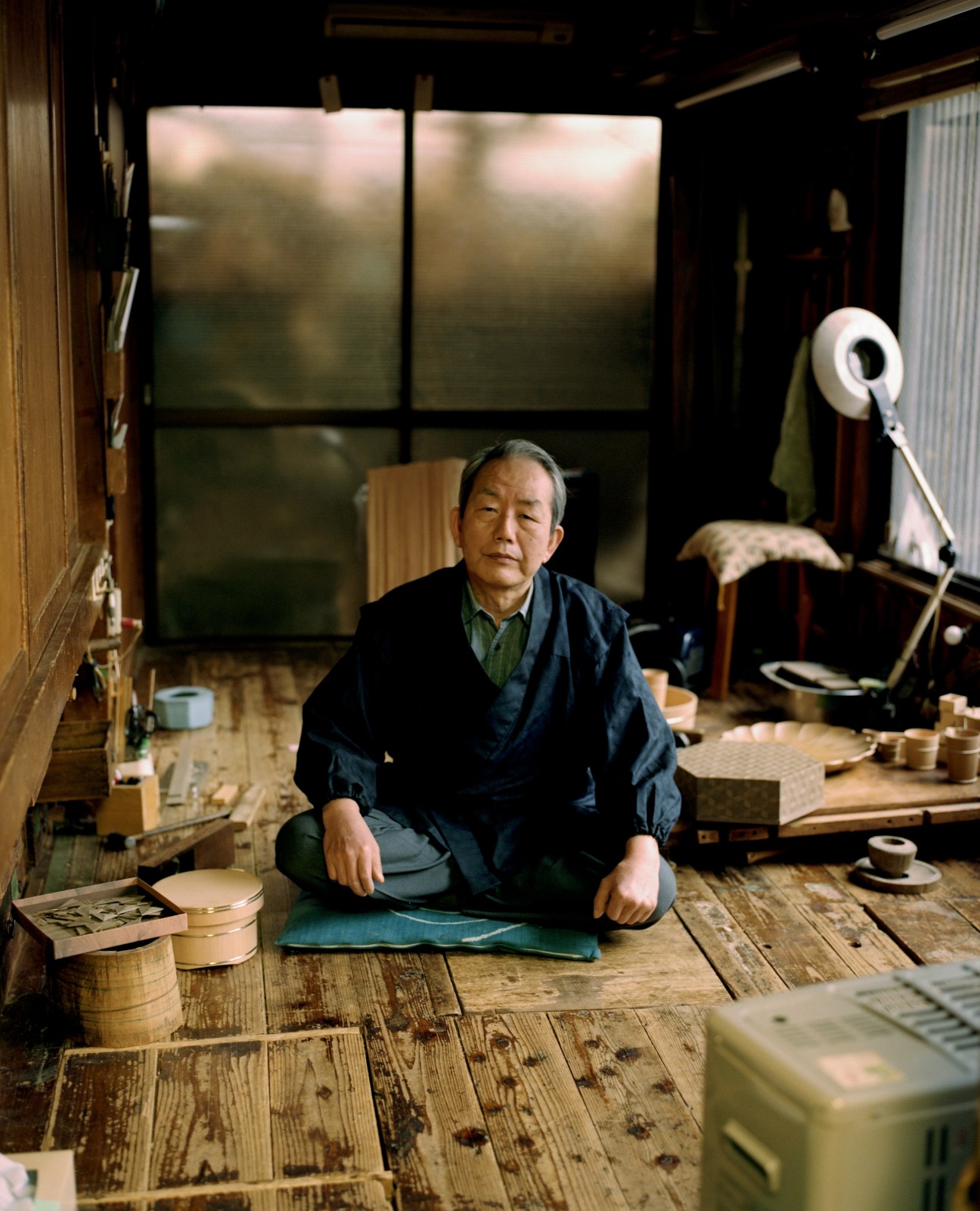 Kiyotsugu Nakagawa, wood artist, sitting in his workshop surrounded by wooden pieces and tools.