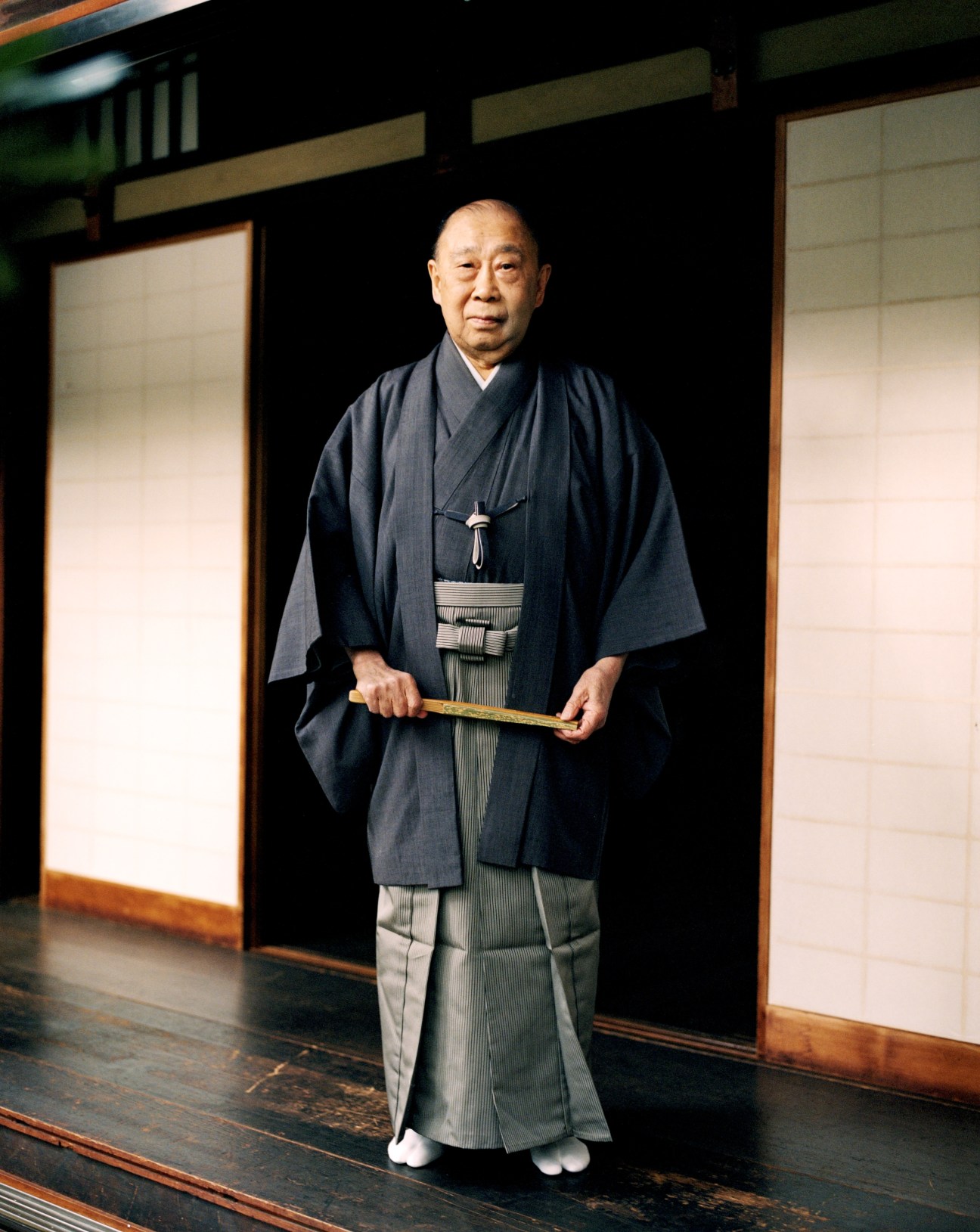 Yusetsu Katayama, a noh performer in his 80s, wearing traditional dark indigo kimono and hakama, holds a wooden fan while standing in a traditional Japanese room.