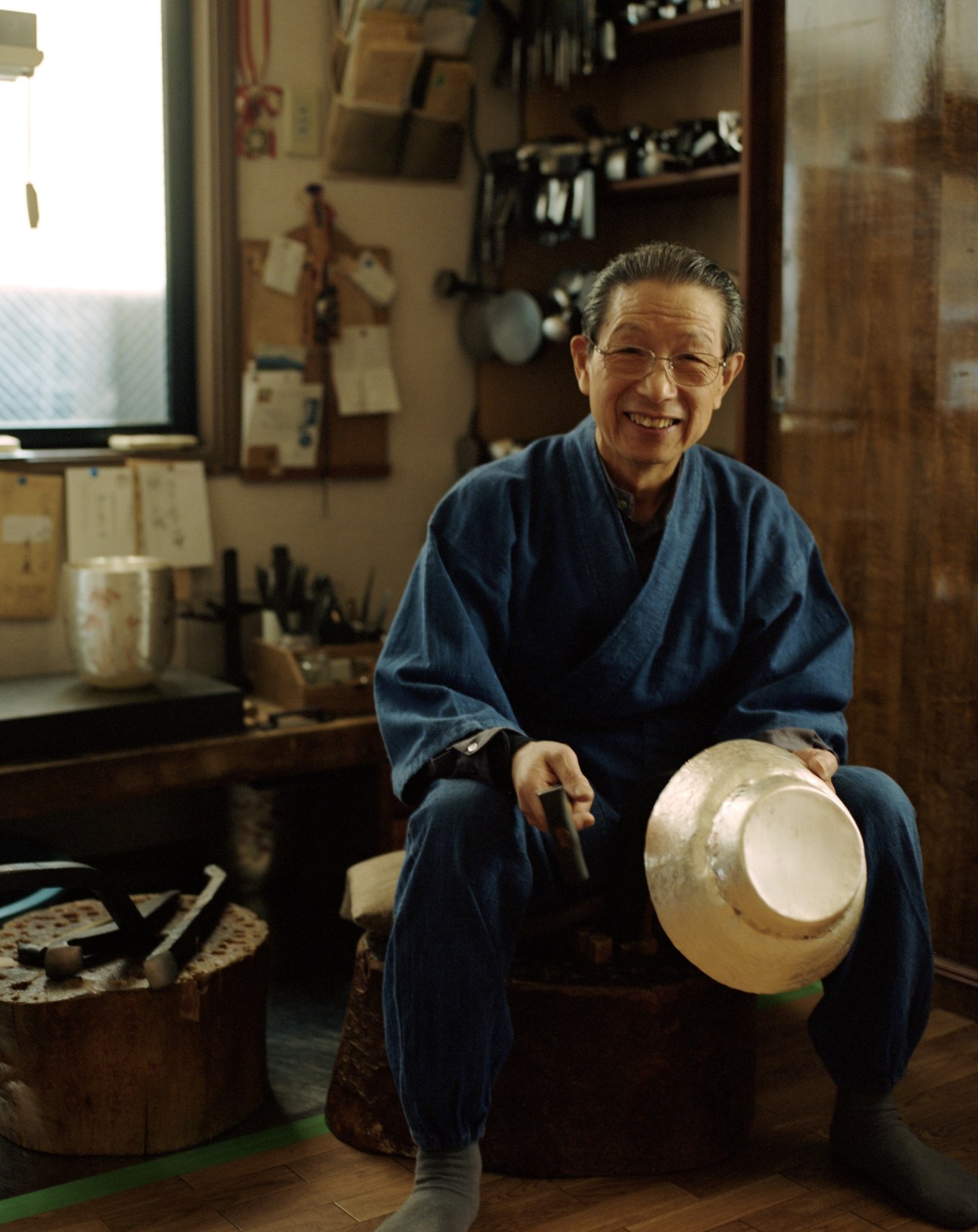 Hoseki Okuyama, metal hammerer, holding a smoothly crafted metal bowl in his Tokyo workshop surrounded by tools.