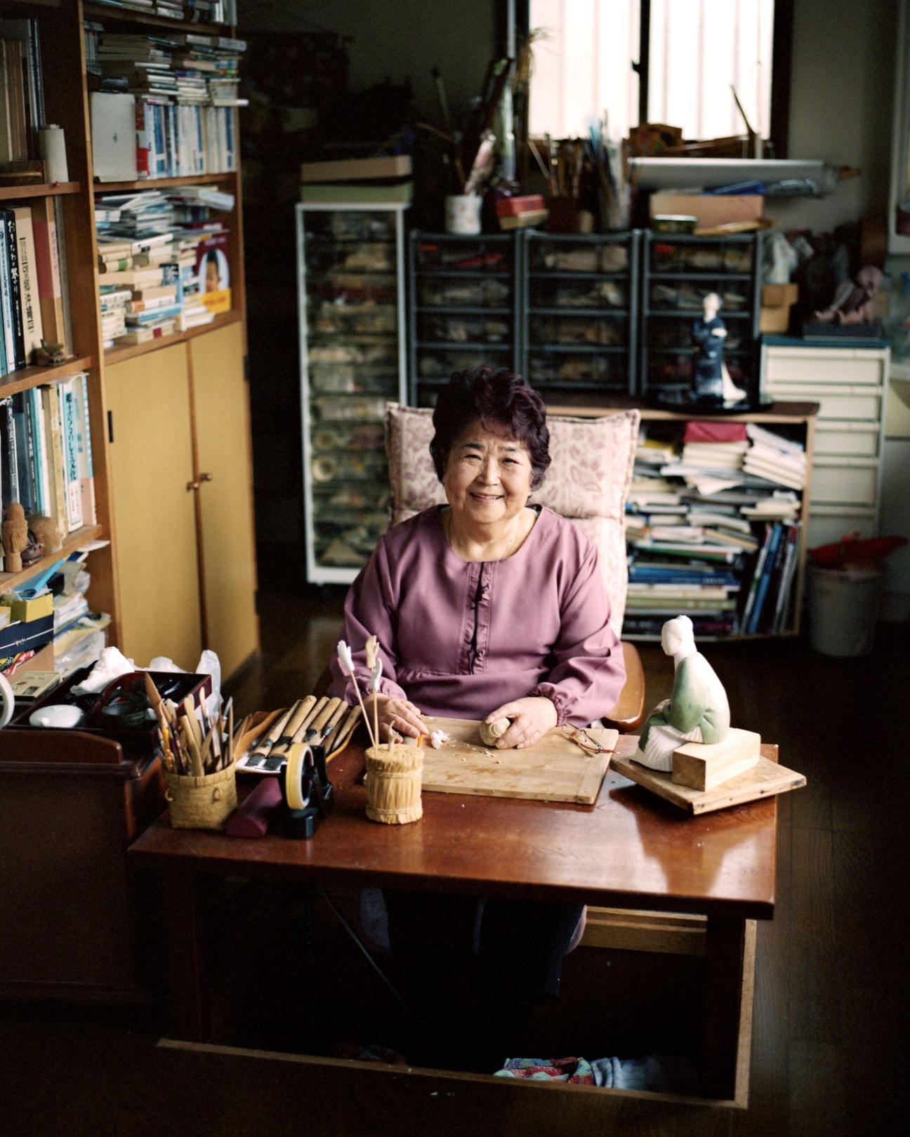 Nobuko Akiyama, Japanese doll maker, sits at her wooden workbench surrounded by carving tools and wooden dolls in her studio.