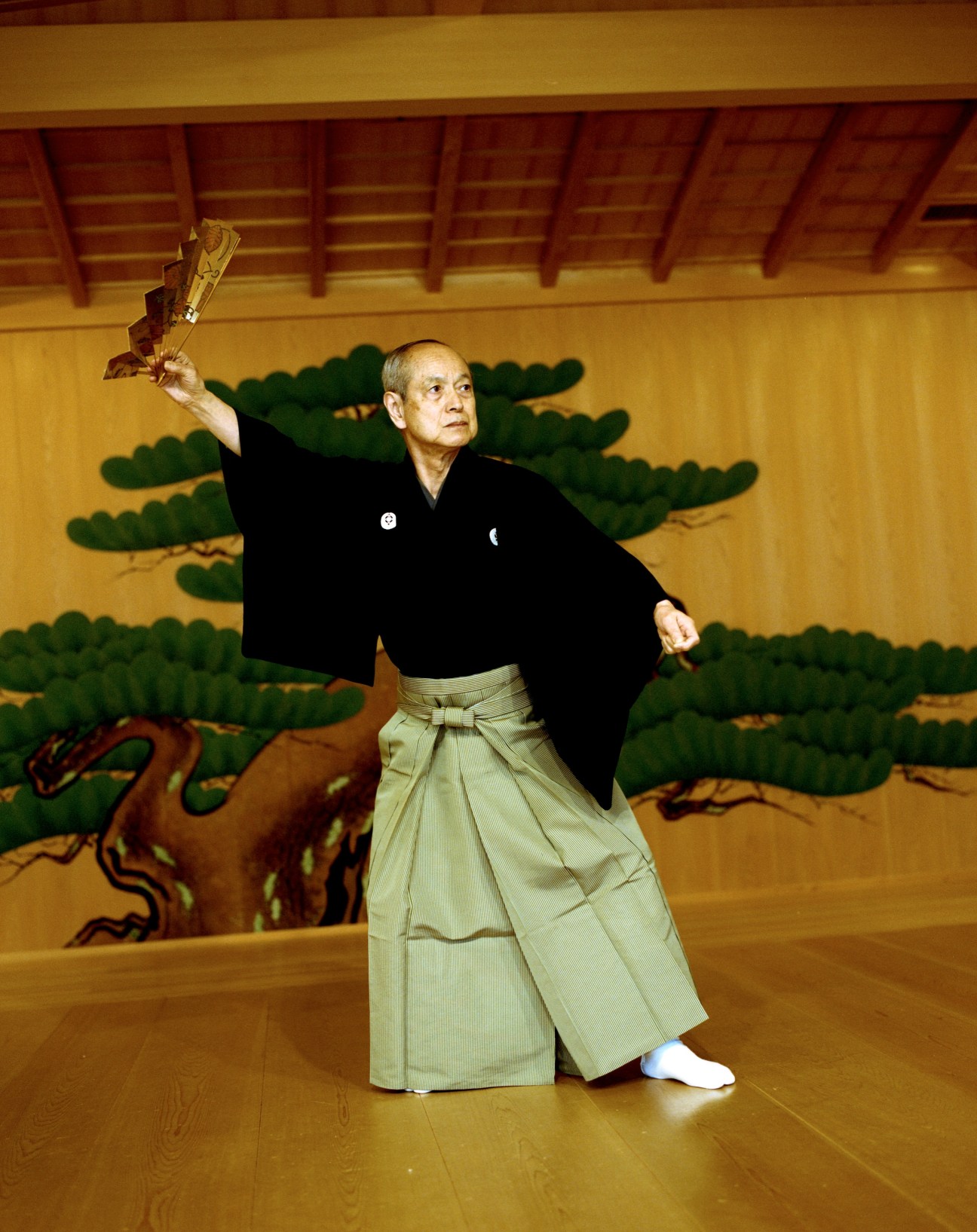 Mansaku Nomura, an elderly kyogen performer in traditional black jacket and light-colored hakama pants, holds a fan aloft on a wooden stage with painted backdrop.