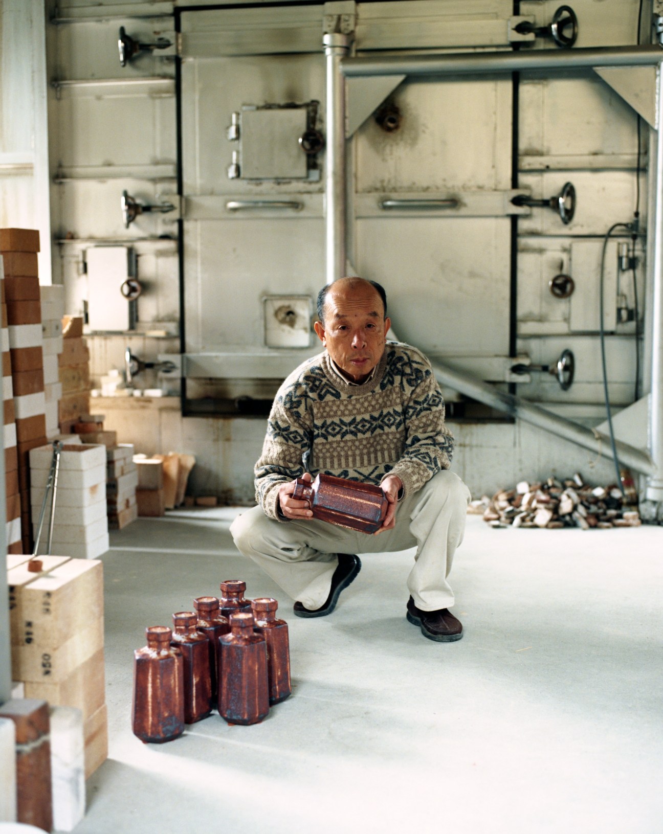 Osamu Suzuki, ceramic artist, crouches in his workshop holding a ceramic vessel surrounded by his work.