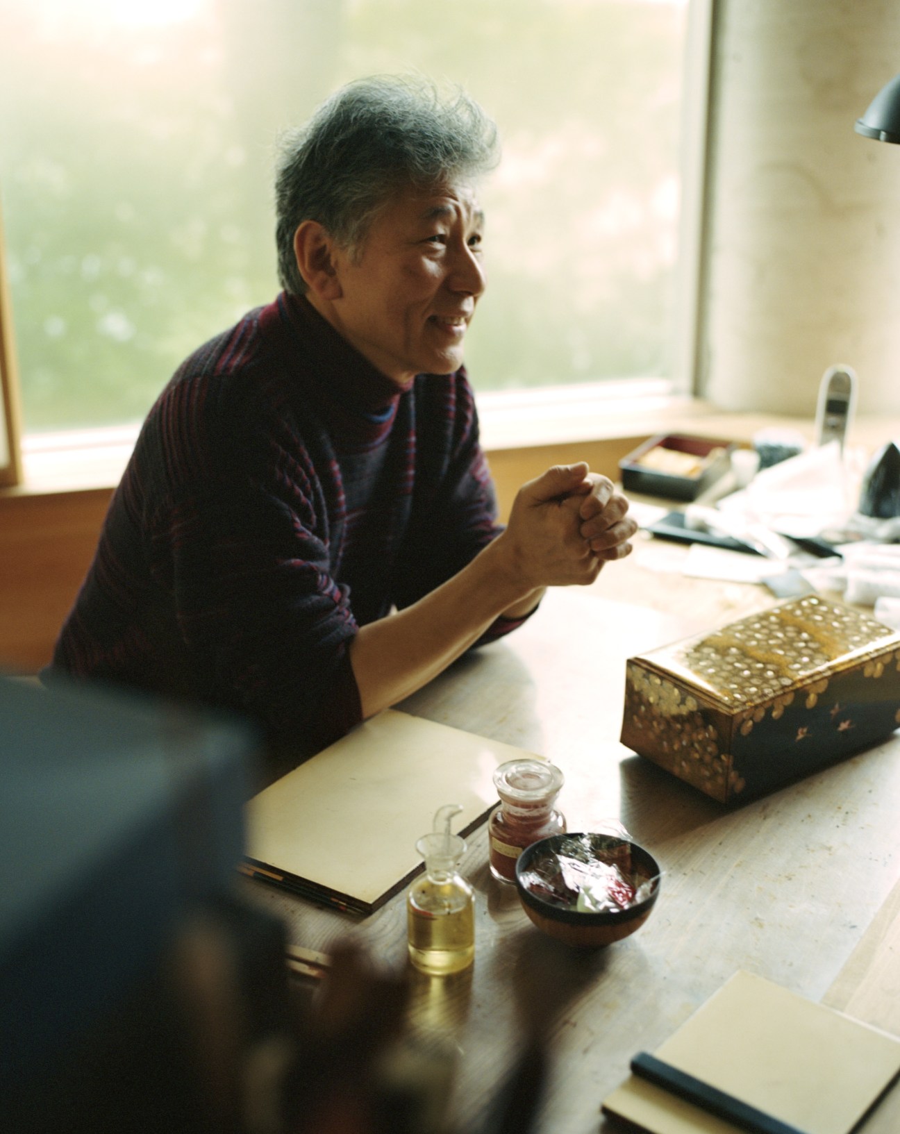 Kazumi Murose, urushi artist, sits at a desk with lacquer work materials including decorative boxes and bottles of lacquer.