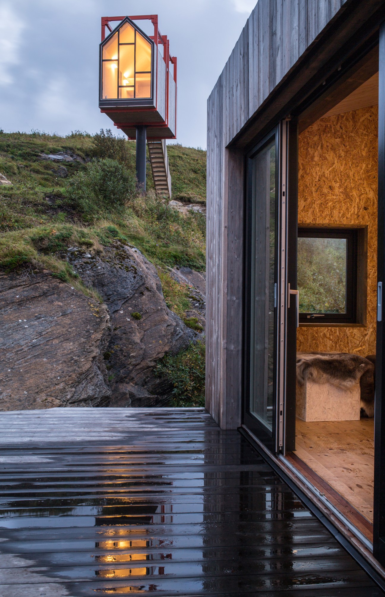 Modern hardwood cabin with illuminated window box on stilts overlooking rocky Norwegian archipelago landscape.