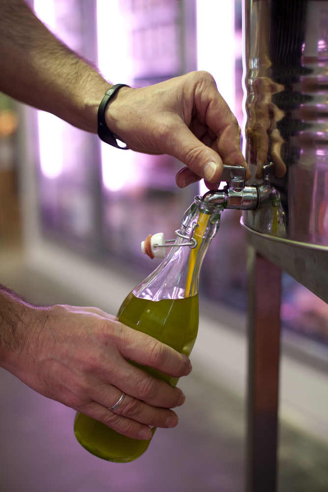 Hand opening a tap to dispense olive oil from Fourth Village's self-service station.