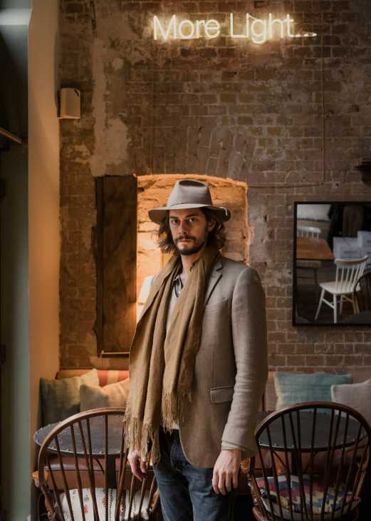 Ludwig Cramer-Klett, founder of Contemporary Food Lab, stands in a brick restaurant interior wearing a hat and scarf.