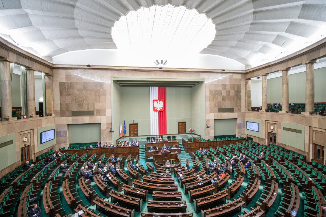 Interior of Polish parliament chamber with green seats, Polish flag, and eagle emblem on wall.