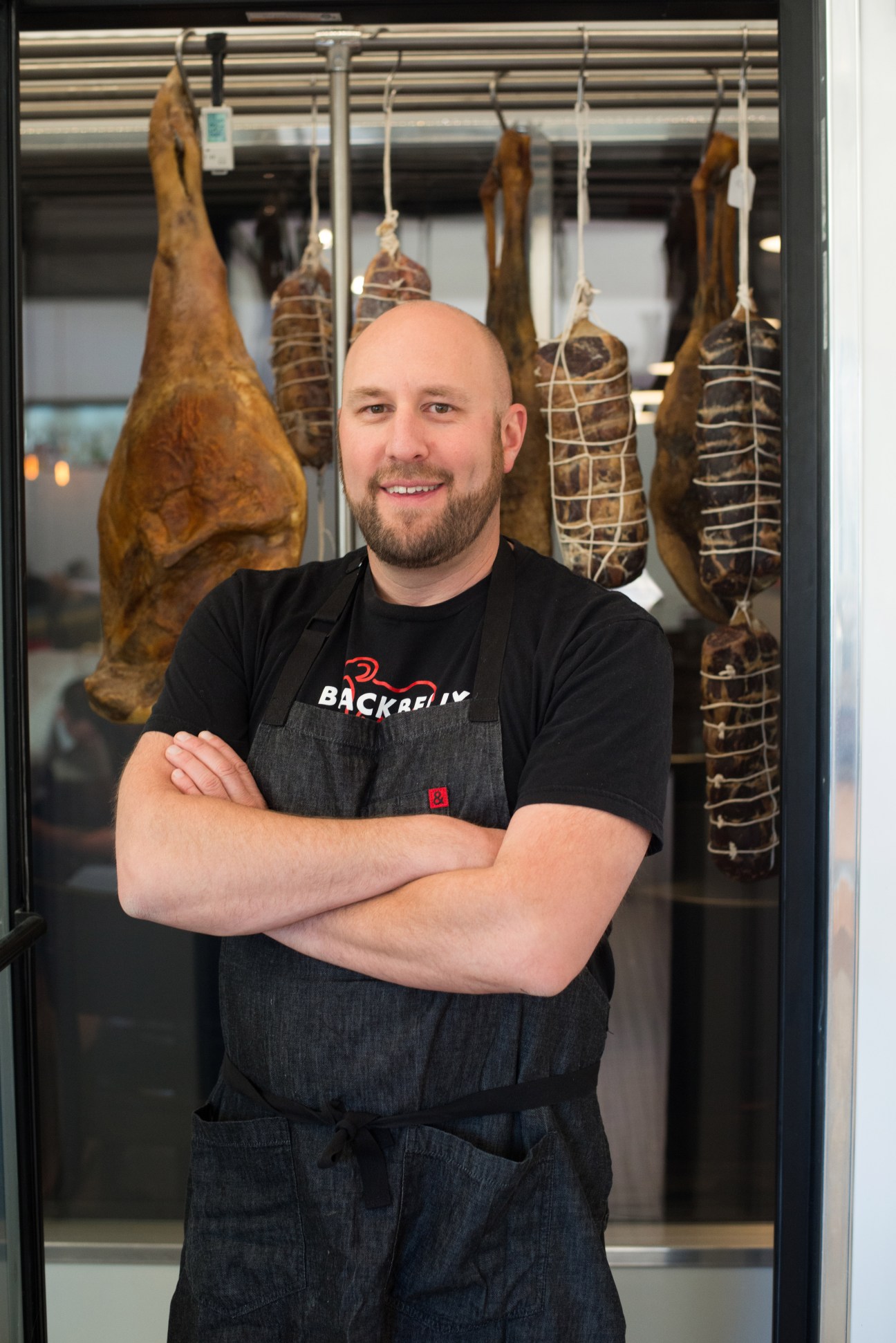 Chef Hosea Rosenberg poses in front of a glass curing chamber with cured meats hanging inside.