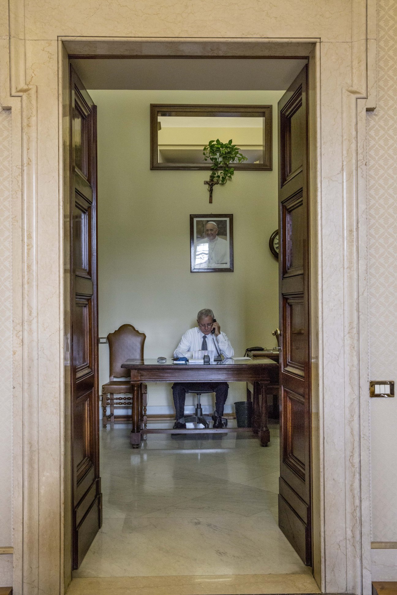 Man in white shirt and tie working at desk in pale green office room with wooden doors and framed portrait on wall.
