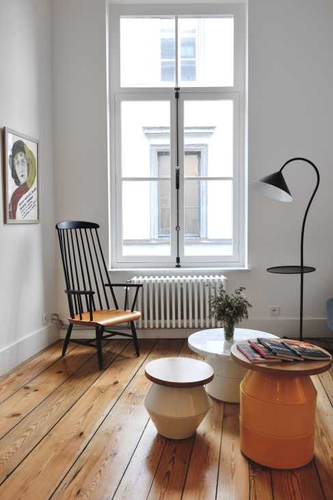 Hotel room interior with wooden chair, side tables, floor lamp, and large windows overlooking street.