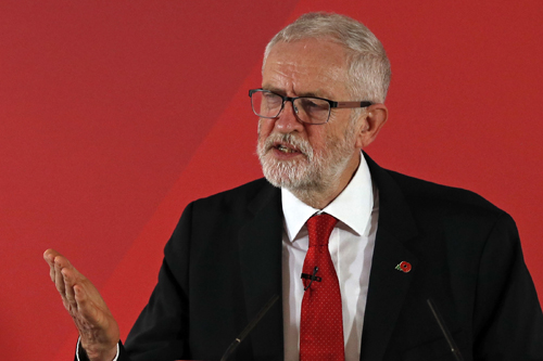 Man in black suit and red tie speaking at a podium against a red background.