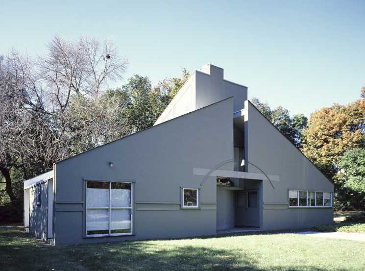 Modern minimalist building with angular geometric forms and large windows surrounded by autumn trees.