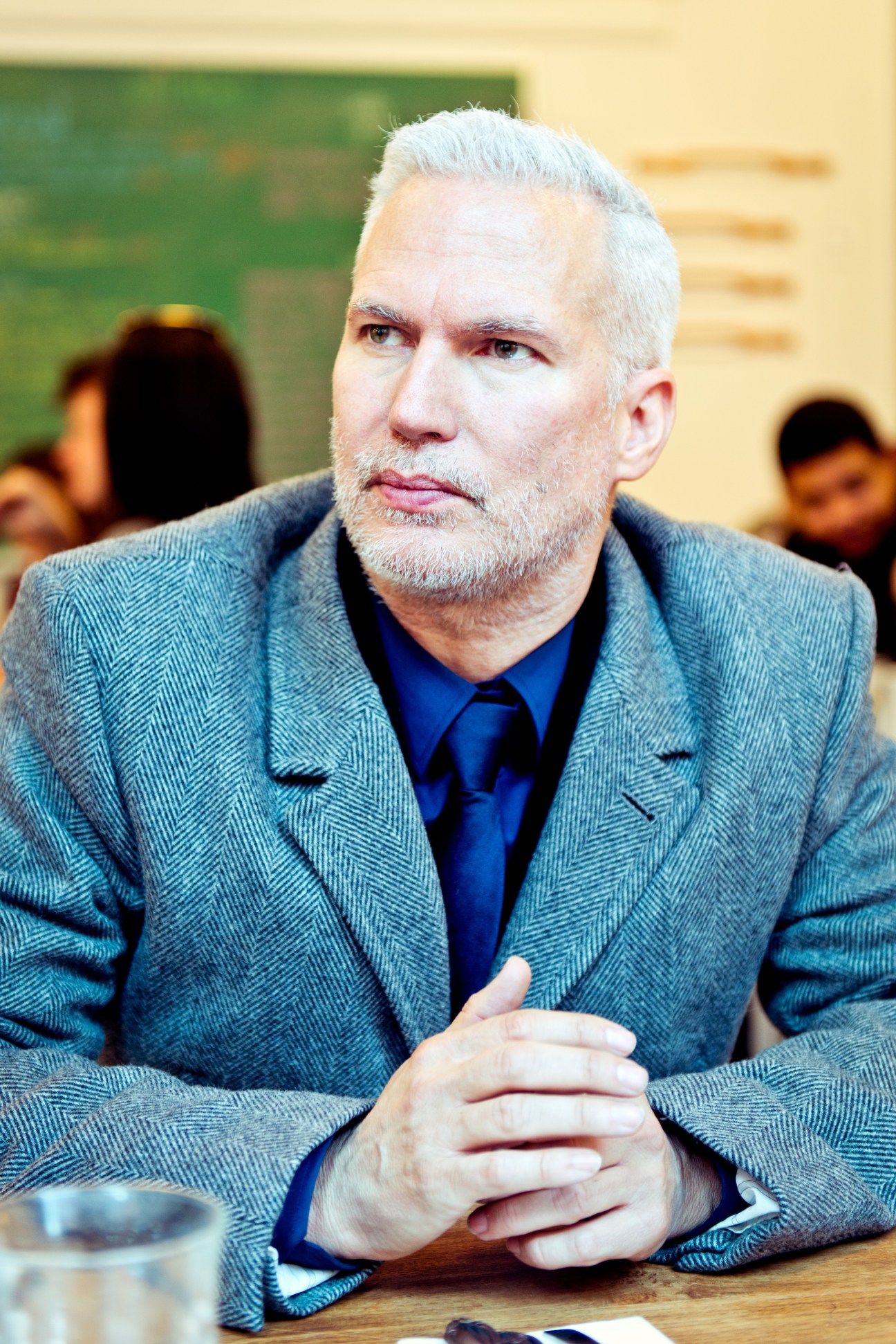 Man with white hair and beard wearing a blue-grey blazer and dark blue shirt at a table.