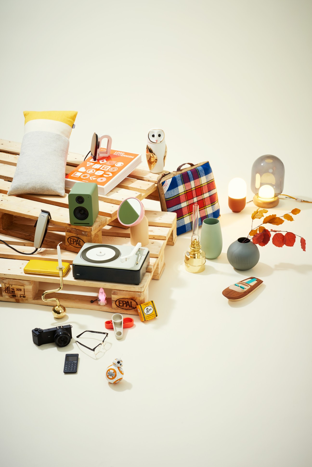 Curated gift items arranged on wooden pallets including speakers, owl vase, lamp, tartan fabric, camera, and Star Wars figure.