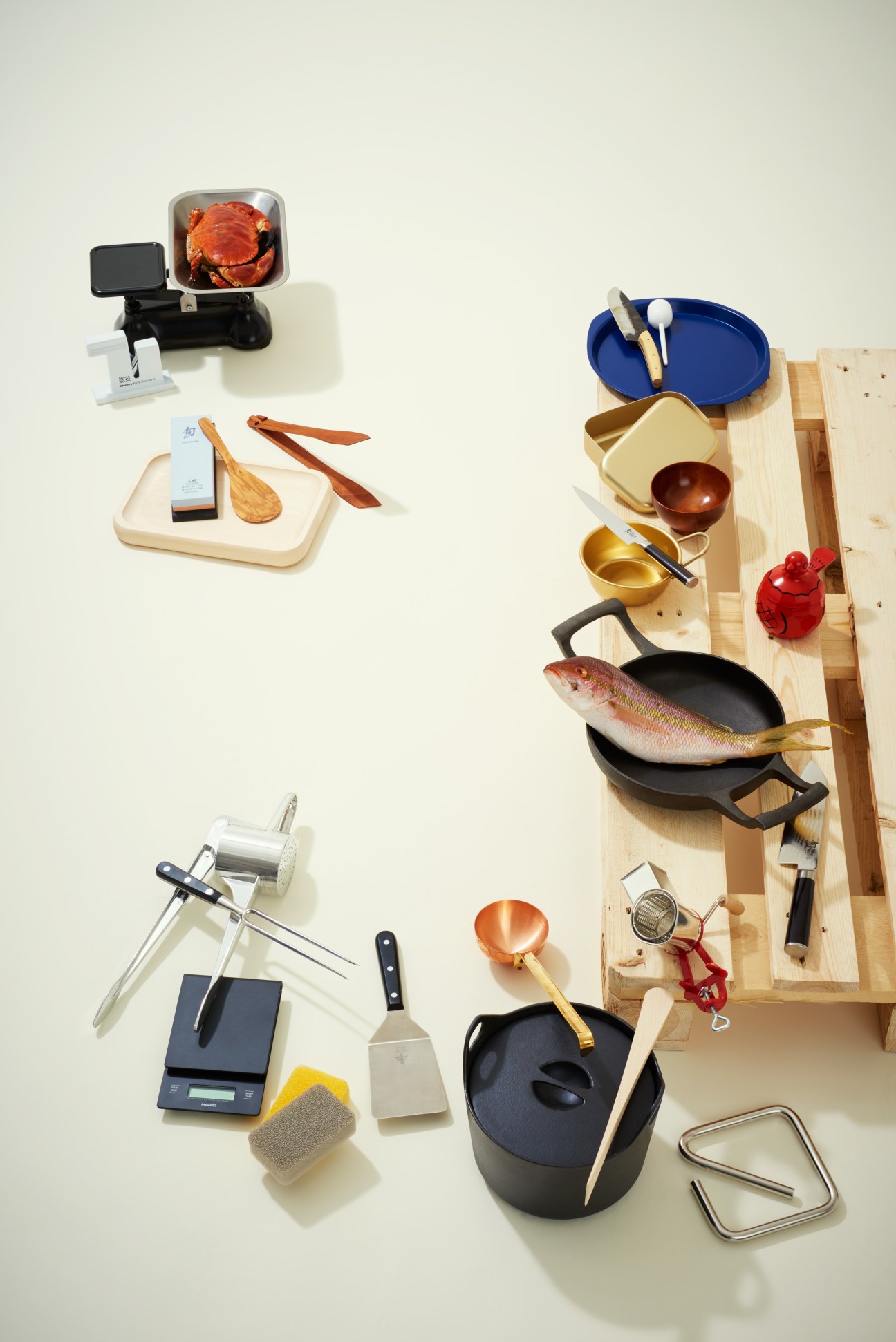 Kitchen tools and cookware arranged on white background including scales, knives, wooden utensils, fish, crab and various cooking implements.