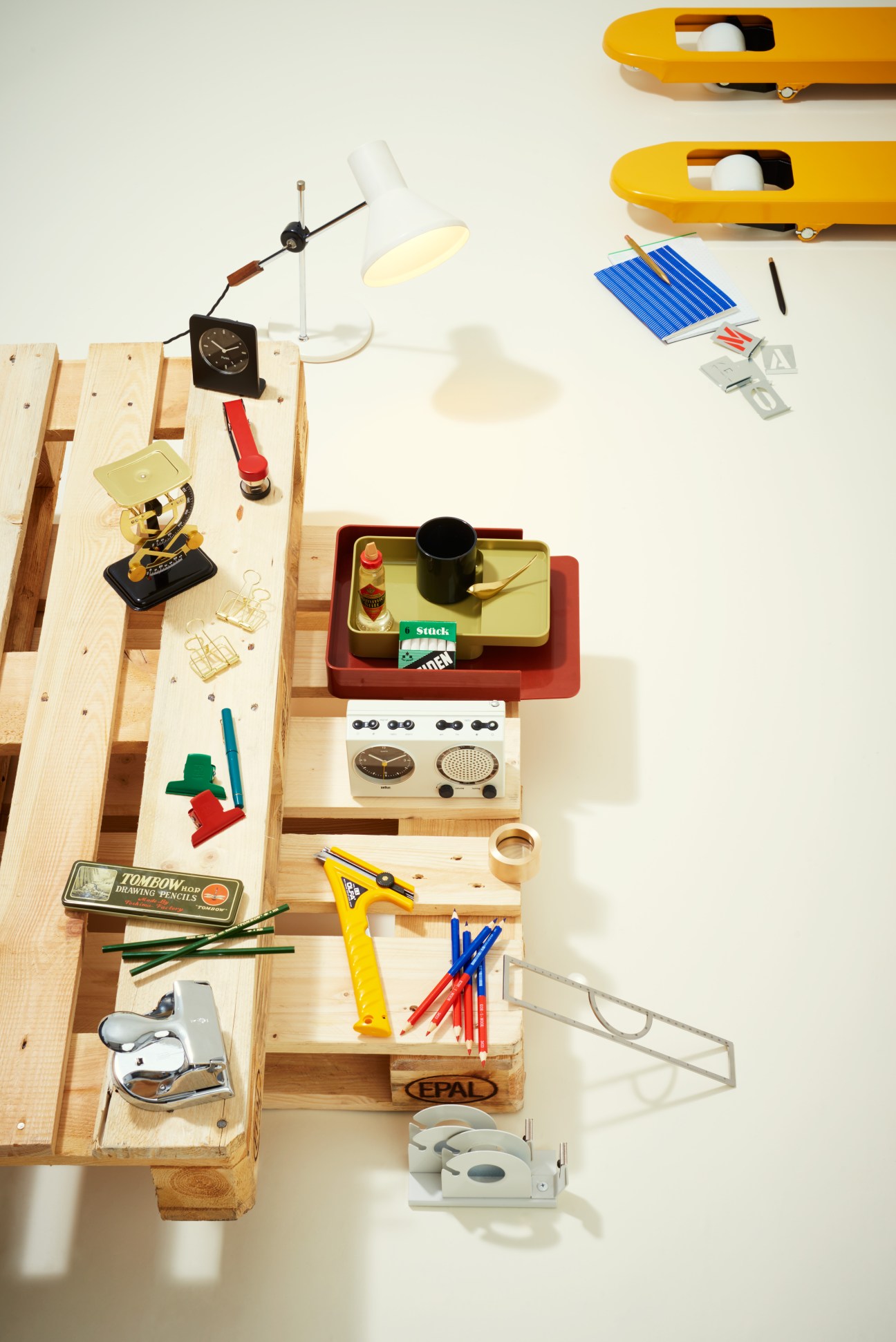 Overhead view of office supplies and stationery arranged on wooden desk organizers, including lamp, clock, stapler, pen, notebook, clips, and scissors.