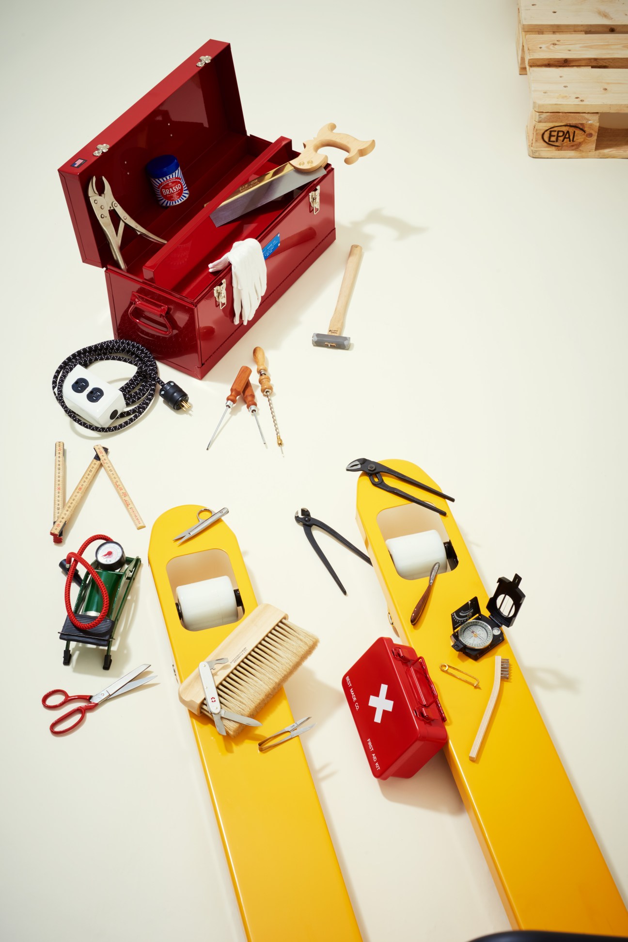 Red toolbox with hand tools arranged on yellow and white background including hammers, screwdrivers, pliers, extension cord, and first-aid kit.