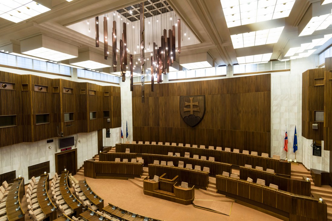 Interior of Slovak parliament chamber with wooden paneling, tiered seating, and a central chandelier.