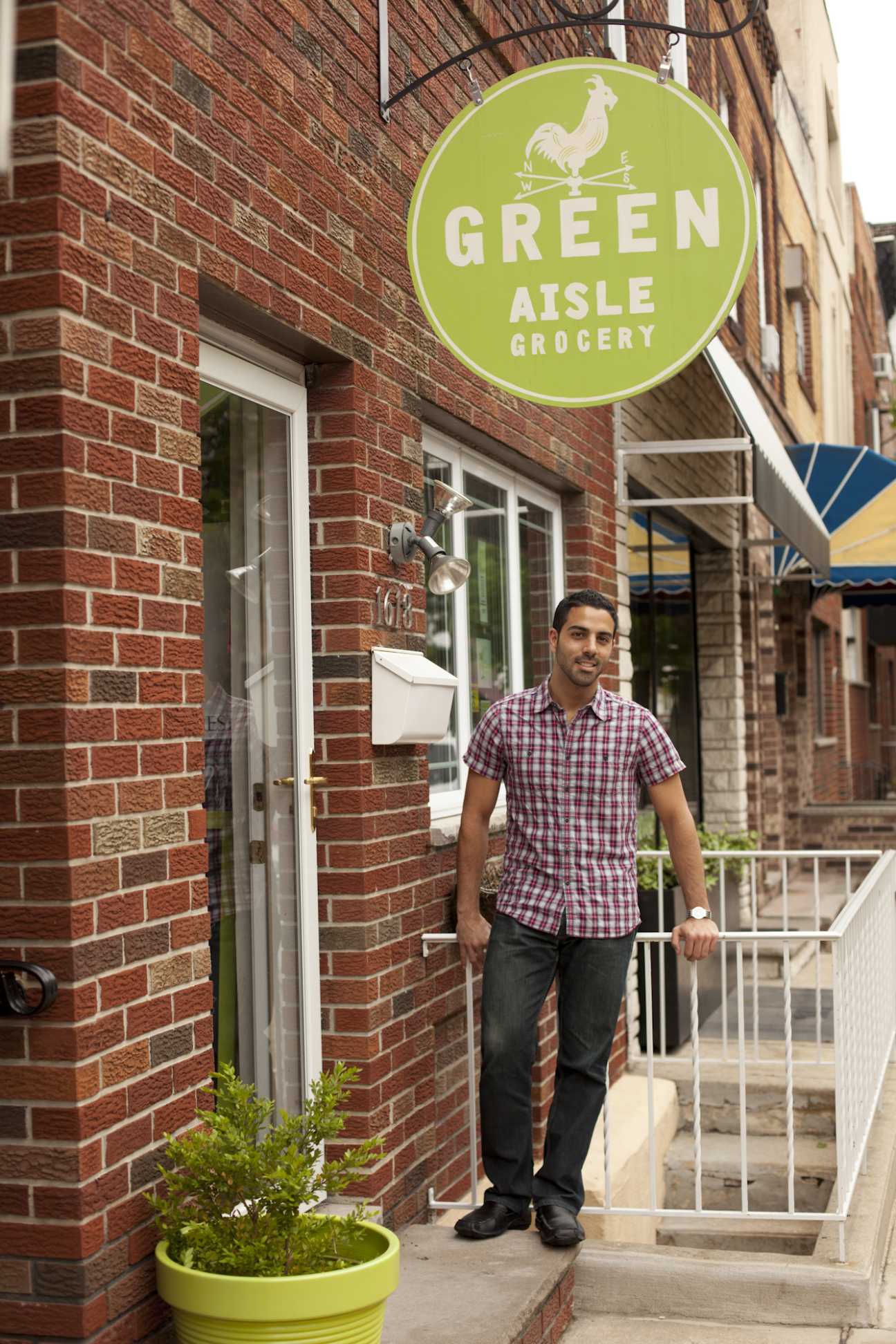 Man standing outside Green Aisle Grocery storefront with lime green circular sign and rooster logo on red brick building.